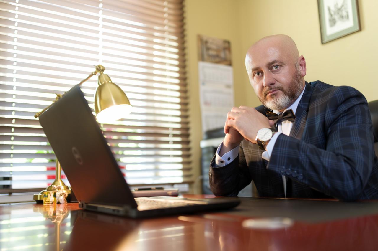 Portret mężczyzny w garniturze i muszce siedzącego za biurkiem z laptopem i lampką, spoglądającego w stronę widza.