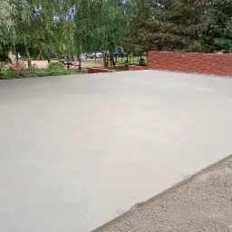 Świeżo wylana, gładka posadzka jastrychowa na zewnątrz, z widocznym fragmentem ceglanego murku i zieleni w tle. Jasna, równa powierzchnia gotowa do dalszych prac wykończeniowych.