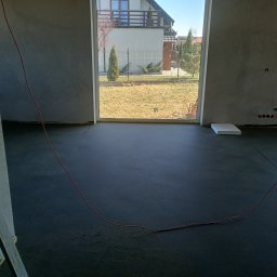 Posadzki Maszynowe Radosław Lisiecki - Gładka, ciemna posadzka w surowym wnętrzu z widokiem na ogród przez duże okno. Czerwony kabel na podłodze. Ściany w stanie deweloperskim.