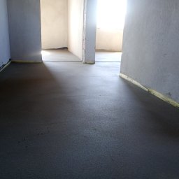 Posadzki Maszynowe Radosław Lisiecki - Świeżo wylana, gładka posadzka w szarym odcieniu, zabezpieczona taśmą przy ścianach. Widok na wnętrze z otwartymi przejściami do kolejnych pomieszczeń.