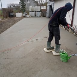 Posadzki Maszynowe Radosław Lisiecki - Pracownik w kaloszach na platformach wyrównuje świeżo wylaną, szarą posadzkę jastrychową na zewnątrz budynku. W tle dom i zbiorniki IBC.