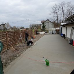 Posadzki Maszynowe Radosław Lisiecki - Wyrównywanie świeżo wylanej, szarej posadzki jastrychowej na zewnątrz budynku. Dwóch pracowników używa poziomicy. W tle ogrodzenie i dom.