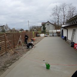 Posadzki Maszynowe Radosław Lisiecki - Dwóch pracowników wyrównuje świeżo wylaną, szarą posadzkę na zewnątrz budynku. W tle widoczny dom, ogrodzenie i drabina oparta o ścianę.