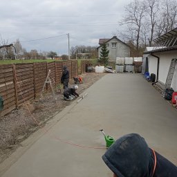Posadzki Maszynowe Radosław Lisiecki - Wyrównywanie świeżo wylanej, szarej posadzki na zewnątrz budynku. Dwóch pracowników w kombinezonach używa poziomicy. W tle widoczny dom i ogrodzenie.
