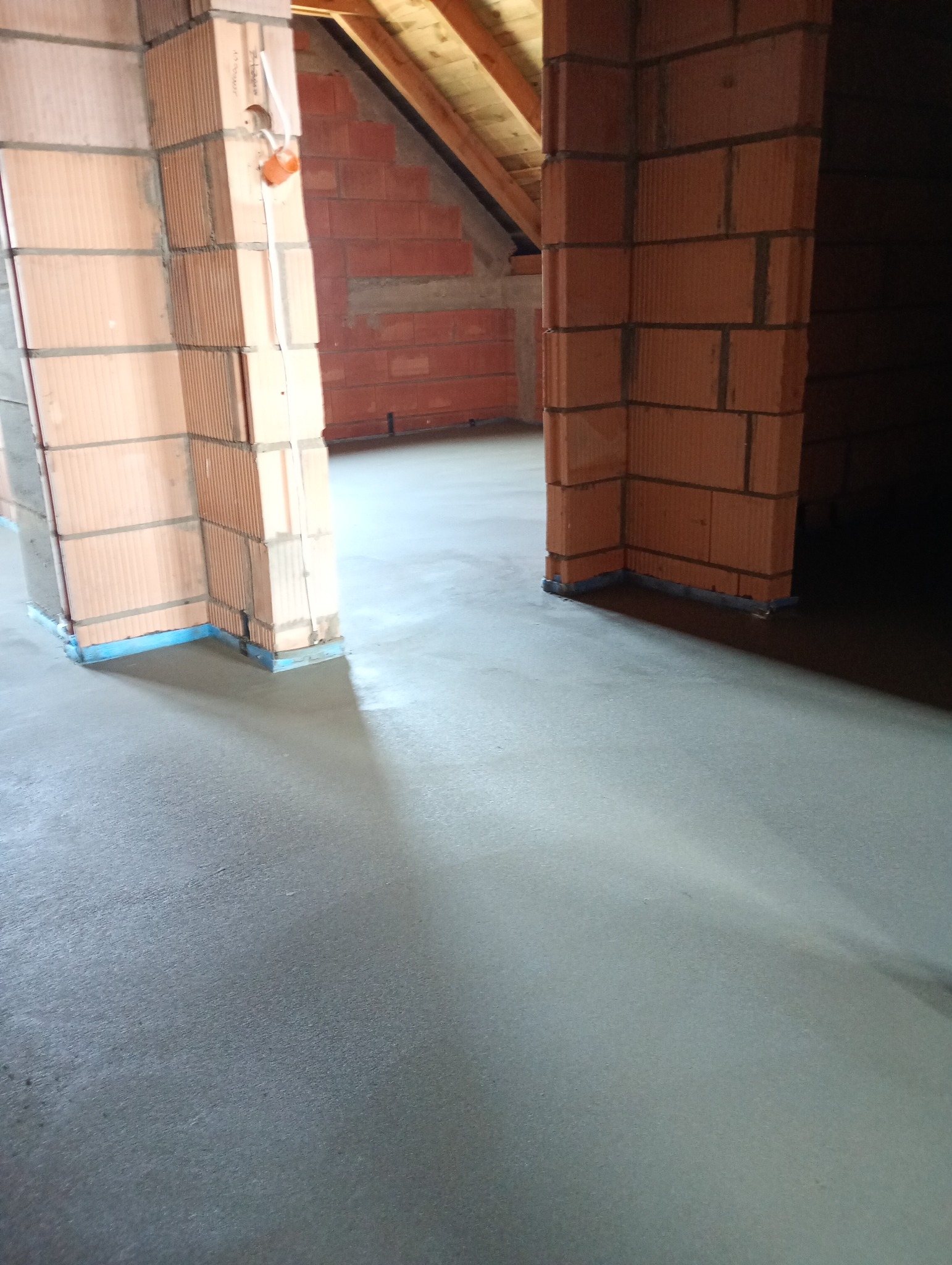 Świeżo wylana, gładka posadzka w surowym stanie, widoczne ceglane filary z niebieską taśmą ochronną przy podłodze, surowa ściana z cegieł w tle.