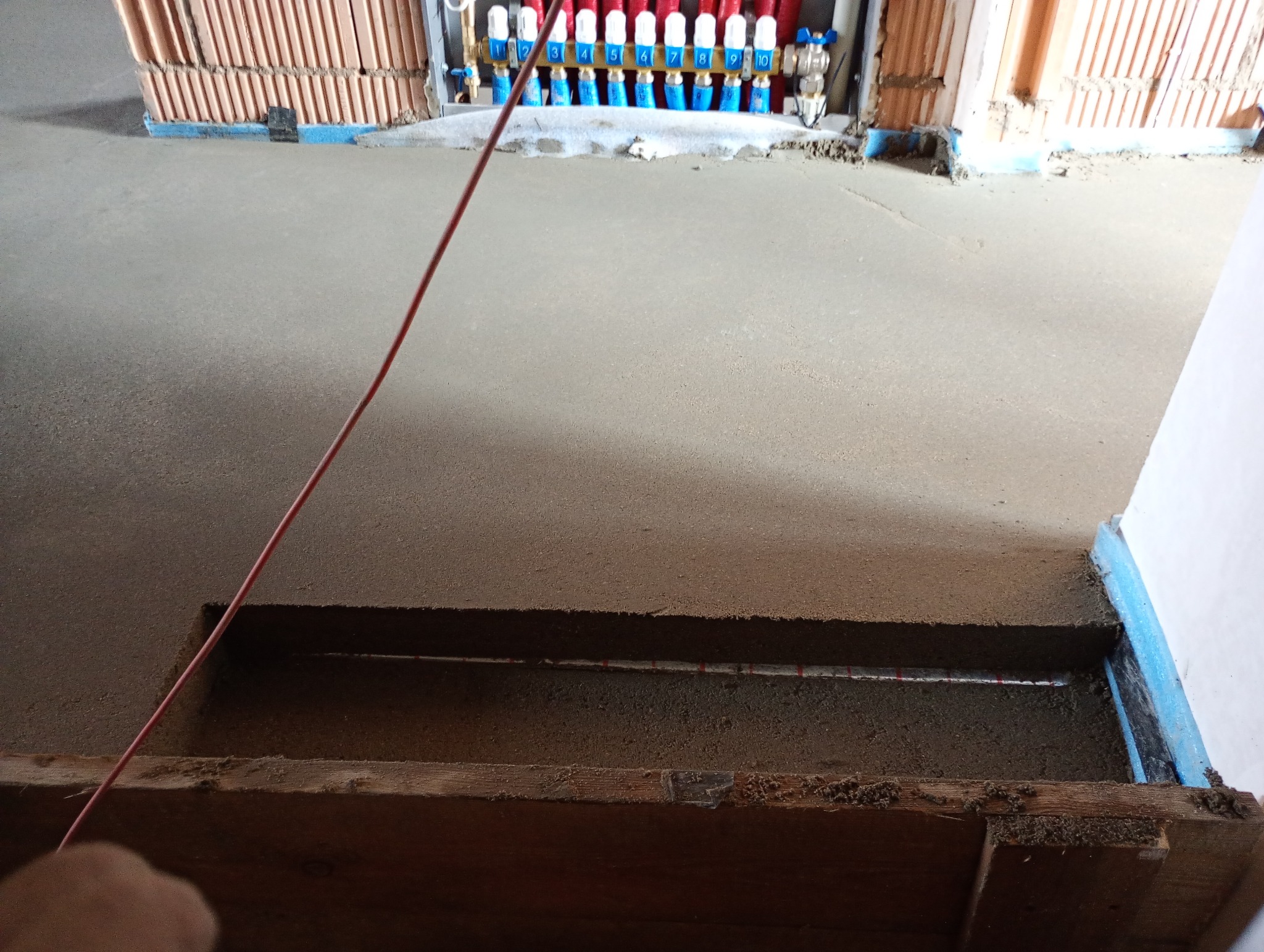 Świeżo wylana, szara posadzka z widocznymi elementami instalacji ogrzewania podłogowego i rozdzielaczem na ścianie w tle.