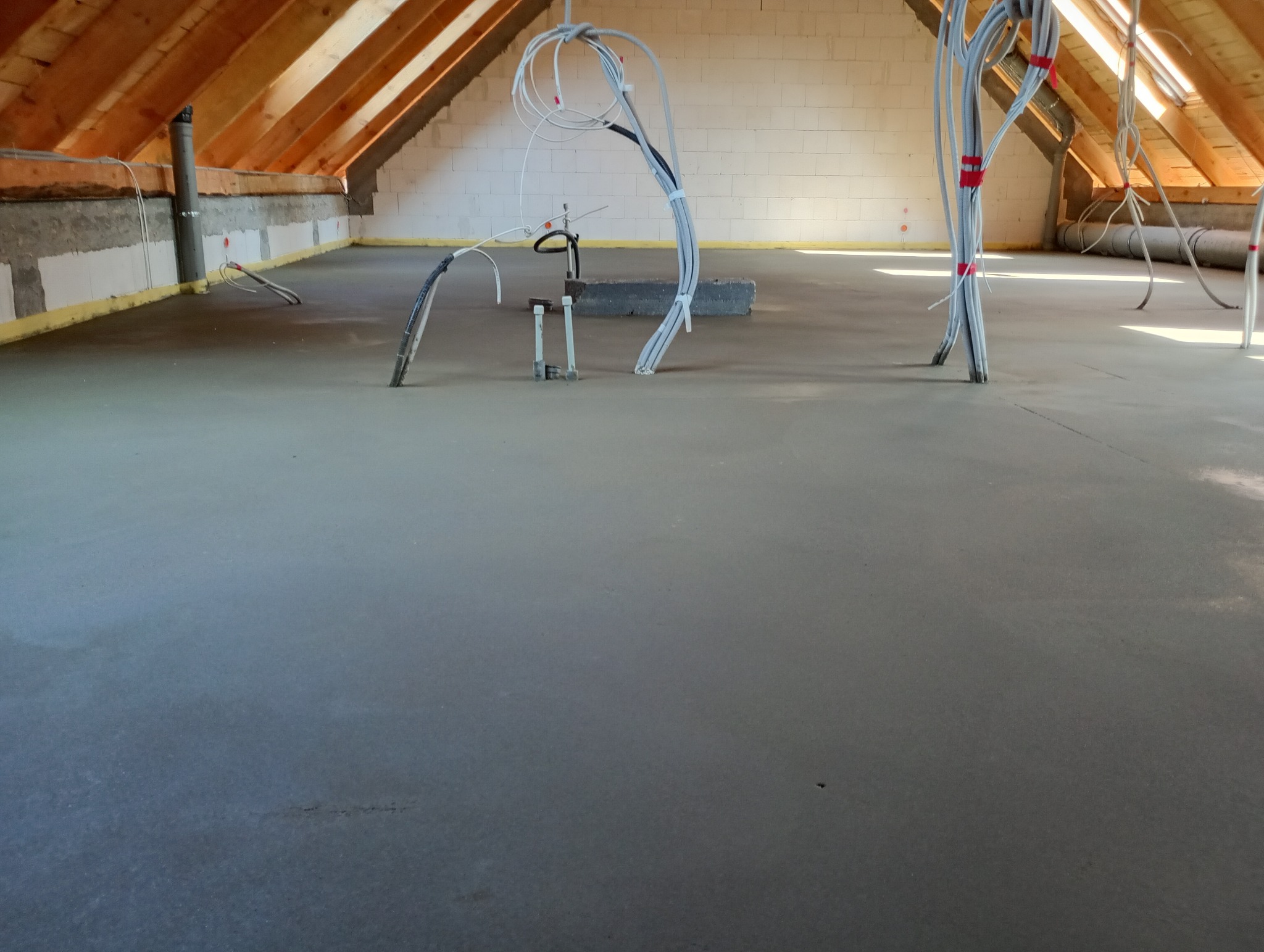 Świeżo wylana, gładka wylewka betonowa w Trzęśniewie Małym na poddaszu, z wystającymi przewodami elektrycznymi i instalacjami, przygotowana pod dalsze prace wykończeniowe.