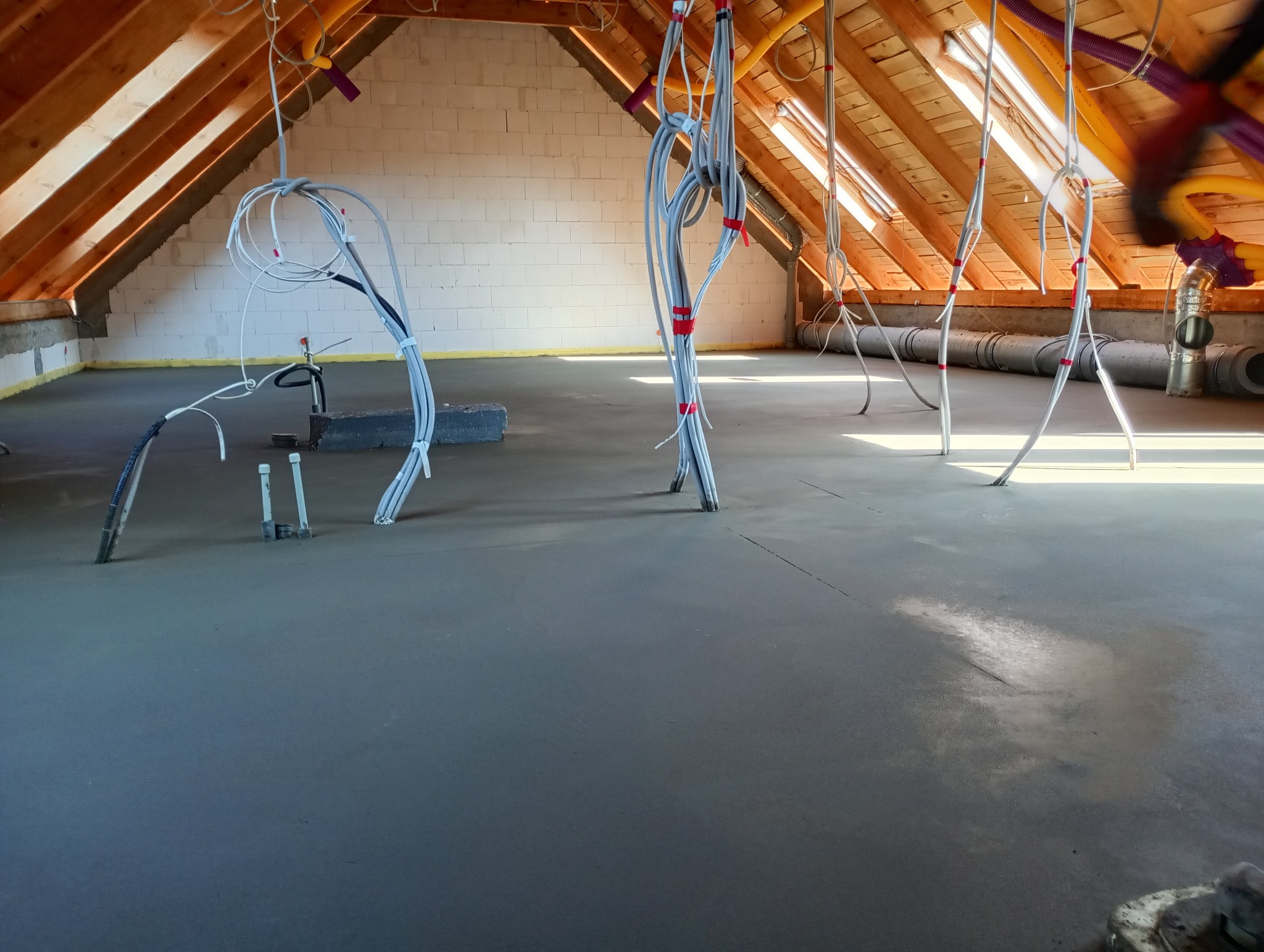 Świeżo wylana, gładka posadzka w poddaszu z widocznymi przewodami instalacji elektrycznej i wentylacyjnej wystającymi z podłoża, drewnianą konstrukcją dachu i ścianami z bloczków.