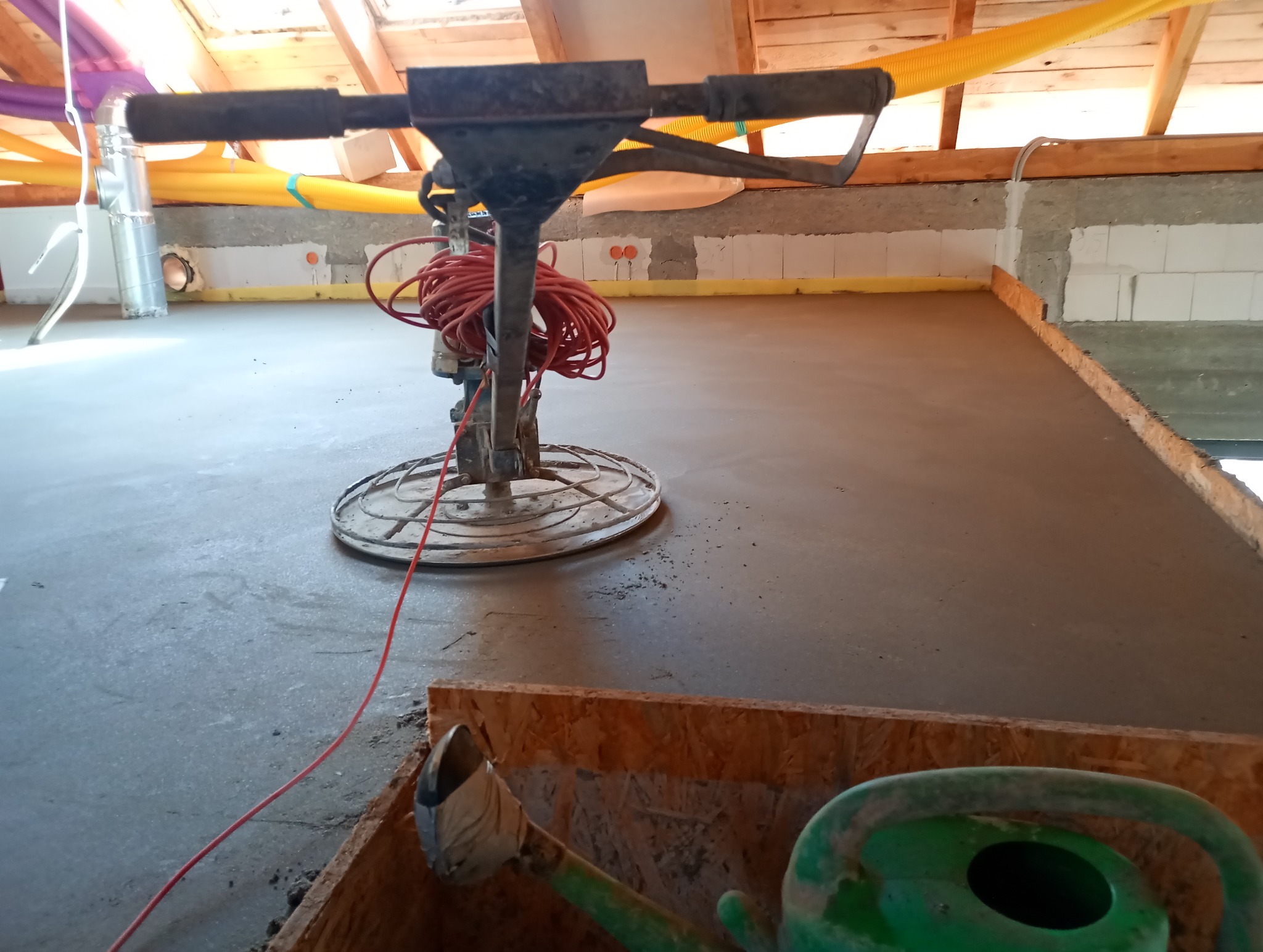 Wyrównywanie wylewki betonowej za pomocą zacieraczki mechanicznej, widoczne instalacje pod sufitem i zielona konewka w rogu kadru.