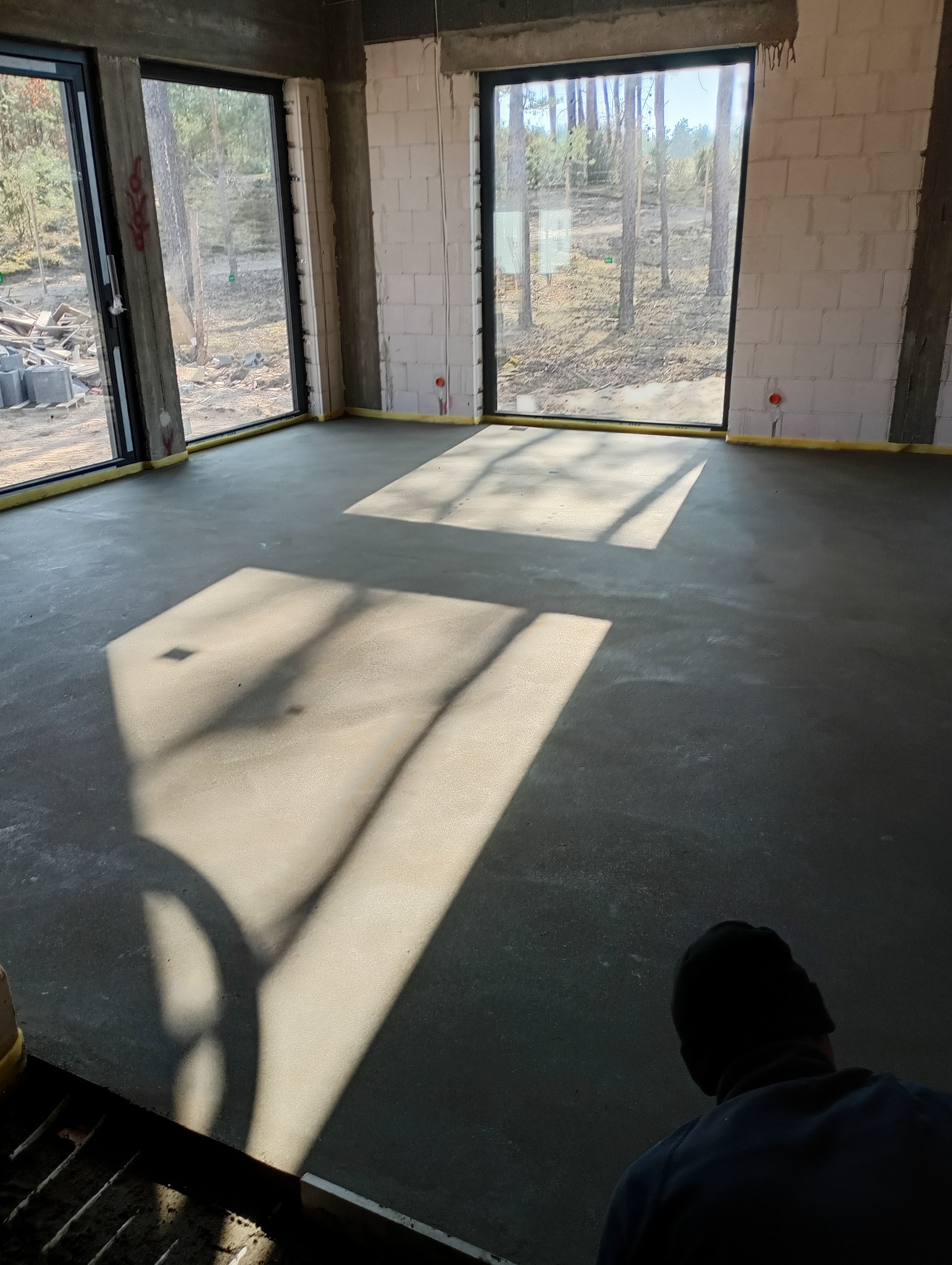 Świeżo wylana, gładka posadzka w surowym wnętrzu domu jednorodzinnego w stanie deweloperskim, z widocznymi promieniami słonecznymi wpadającymi przez duże okna.