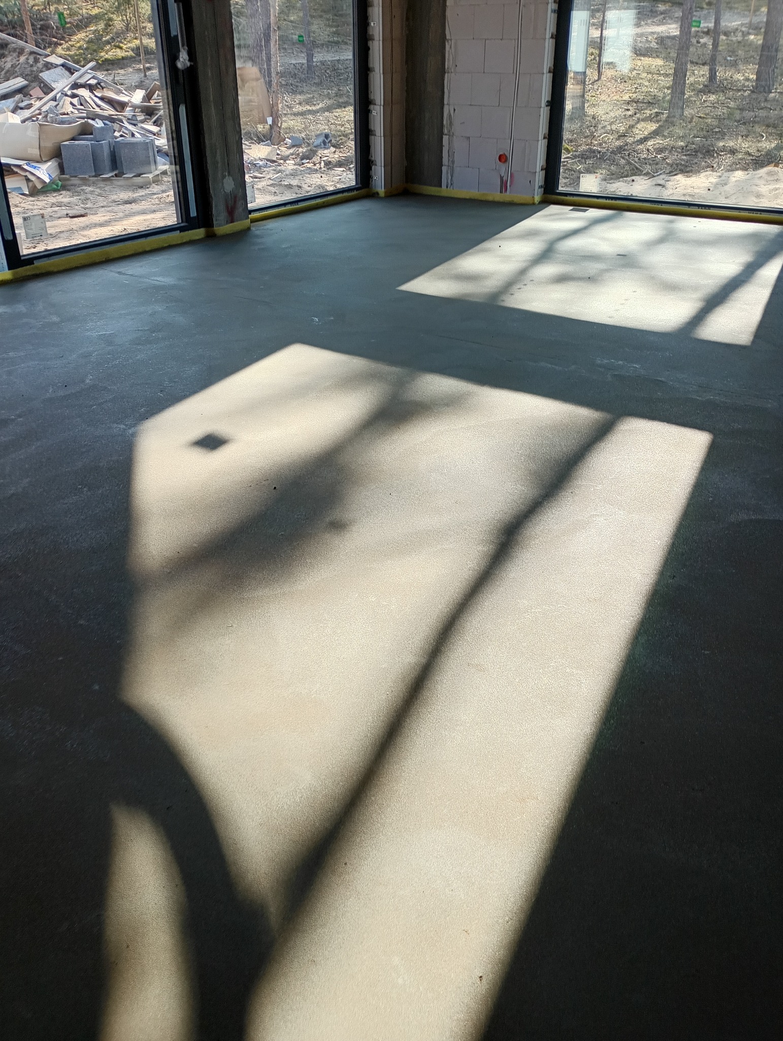 Świeżo wylana, gładka posadzka w surowym wnętrzu budynku w stanie deweloperskim, z widocznymi promieniami słońca wpadającymi przez duże okna.