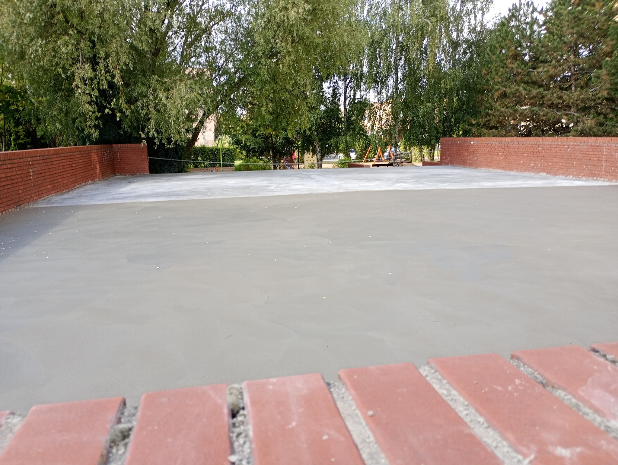 Świeżo wylana, gładka posadzka jastrychowa otoczona niskim, ceglanym murkiem na zewnątrz, z zielenią drzew w tle. Widoczne ślady po narzędziach.