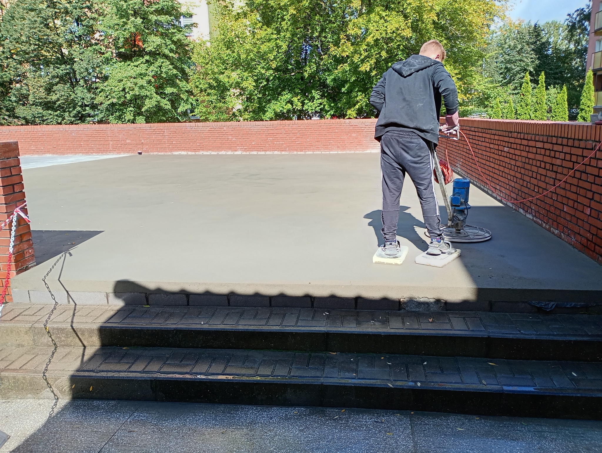 Pracownik w czarnym stroju zaciera świeżą posadzkę jastrychową na zewnątrz, używając specjalistycznej maszyny. Ceglany mur w tle, zielone drzewa.