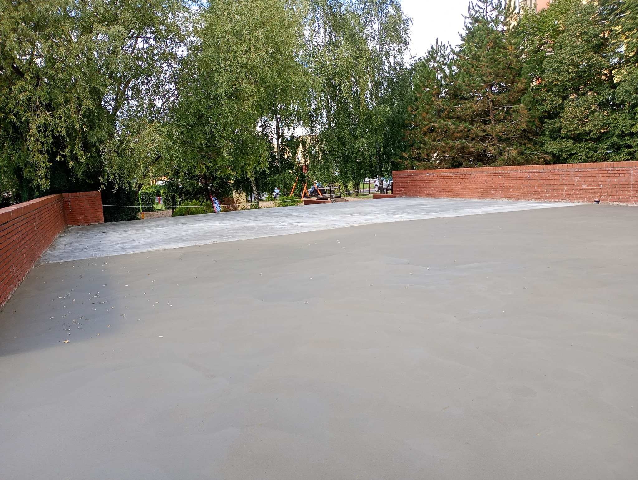 Świeżo wylana, gładka posadzka jastrychowa otoczona niskim, ceglanym murkiem. W tle zieleń drzew i budynki. Widok z góry.