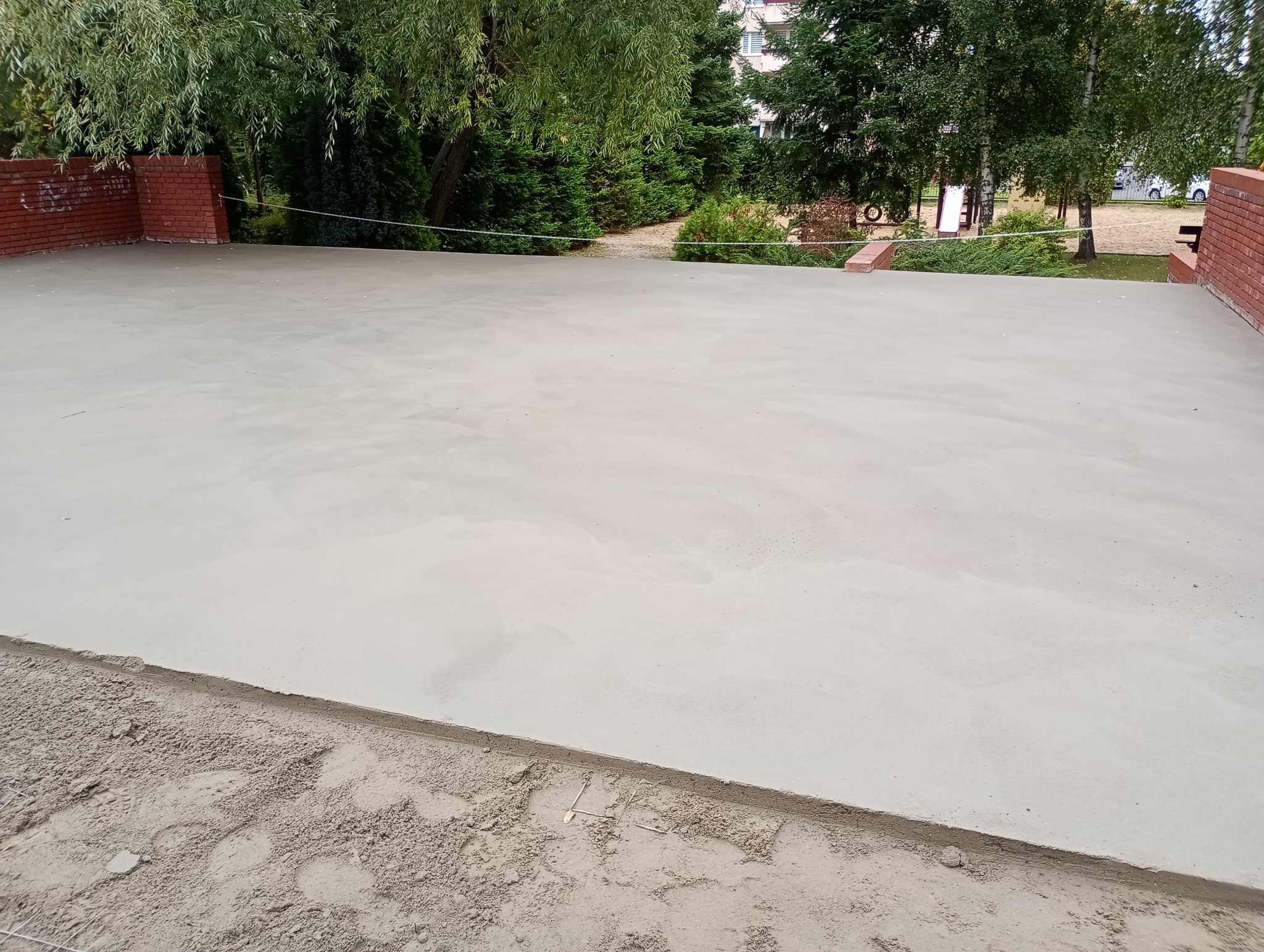 Gładka, szara posadzka jastrychowa na zewnątrz, otoczona ceglanym murkiem i zielenią. Widoczne ślady piasku przy krawędzi.