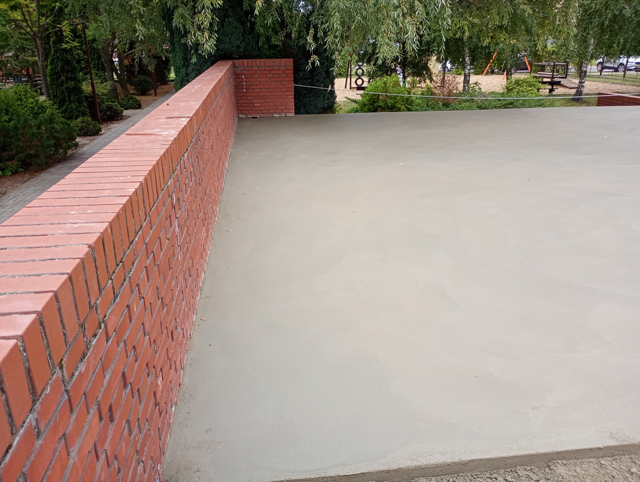 Świeżo wylana, gładka posadzka jastrychowa na zewnątrz, obok mur z czerwonej cegły i zieleń drzew w tle. Widok z góry, jasne oświetlenie.