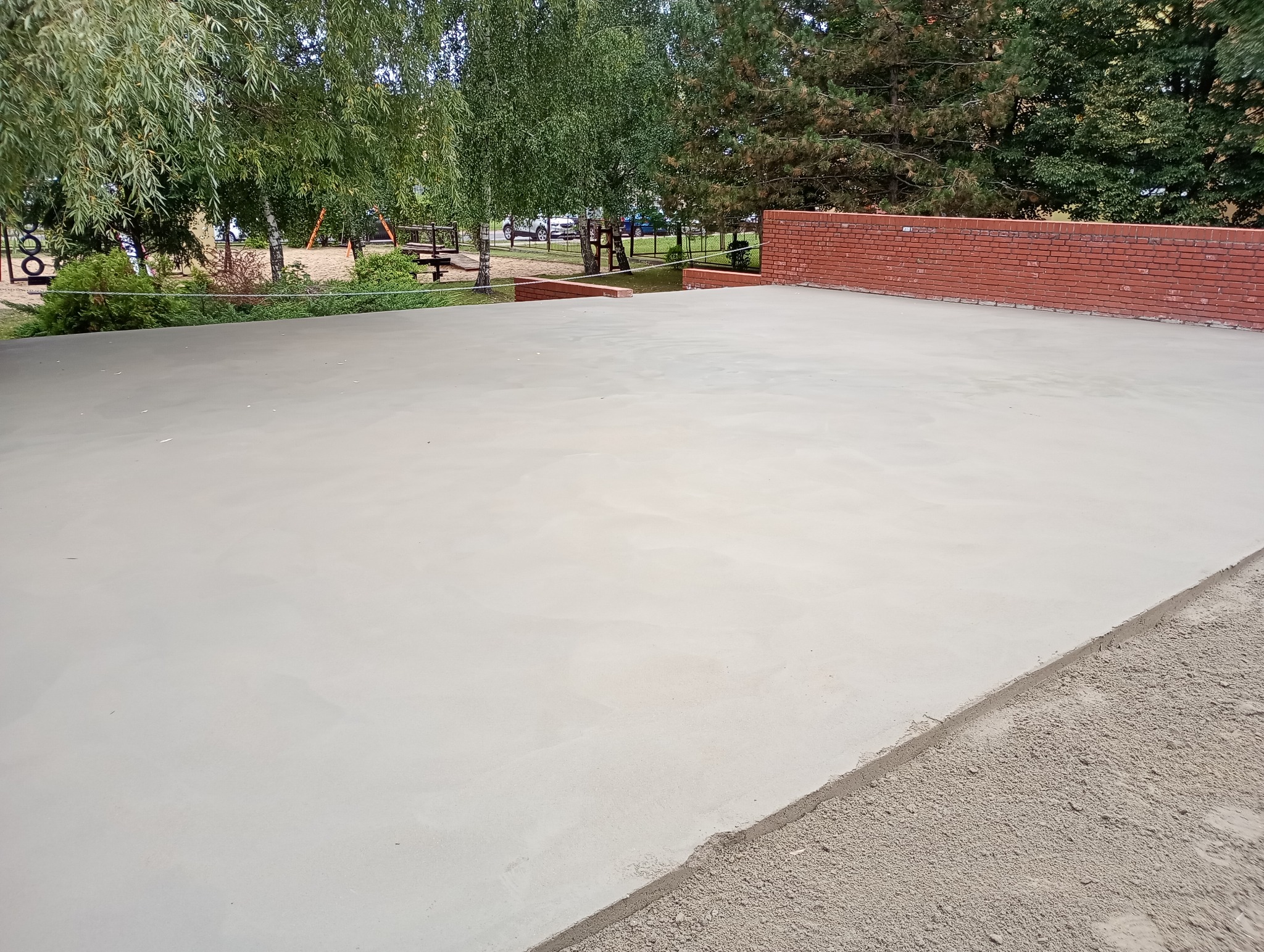 Świeżo wylana, gładka posadzka jastrychowa na zewnątrz, z widocznym fragmentem ceglanego murku i zieleni w tle. Jasna, równa powierzchnia gotowa do dalszych prac wykończeniowych.