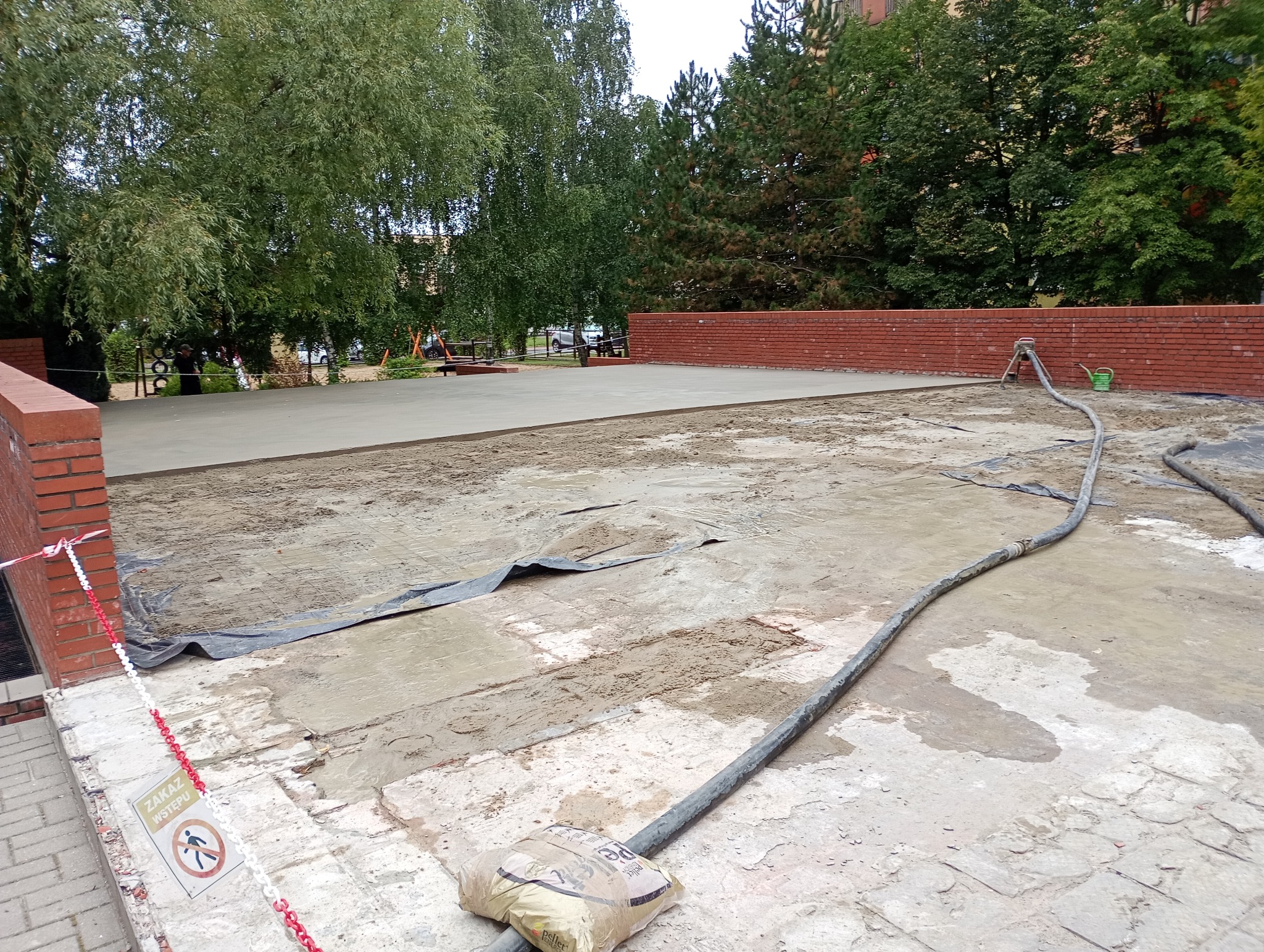 Wylewka jastrychowa na zewnątrz budynku, z widoczną pompą i wężami. Częściowo zalana powierzchnia, z workiem cementu w rogu. Prace w toku.