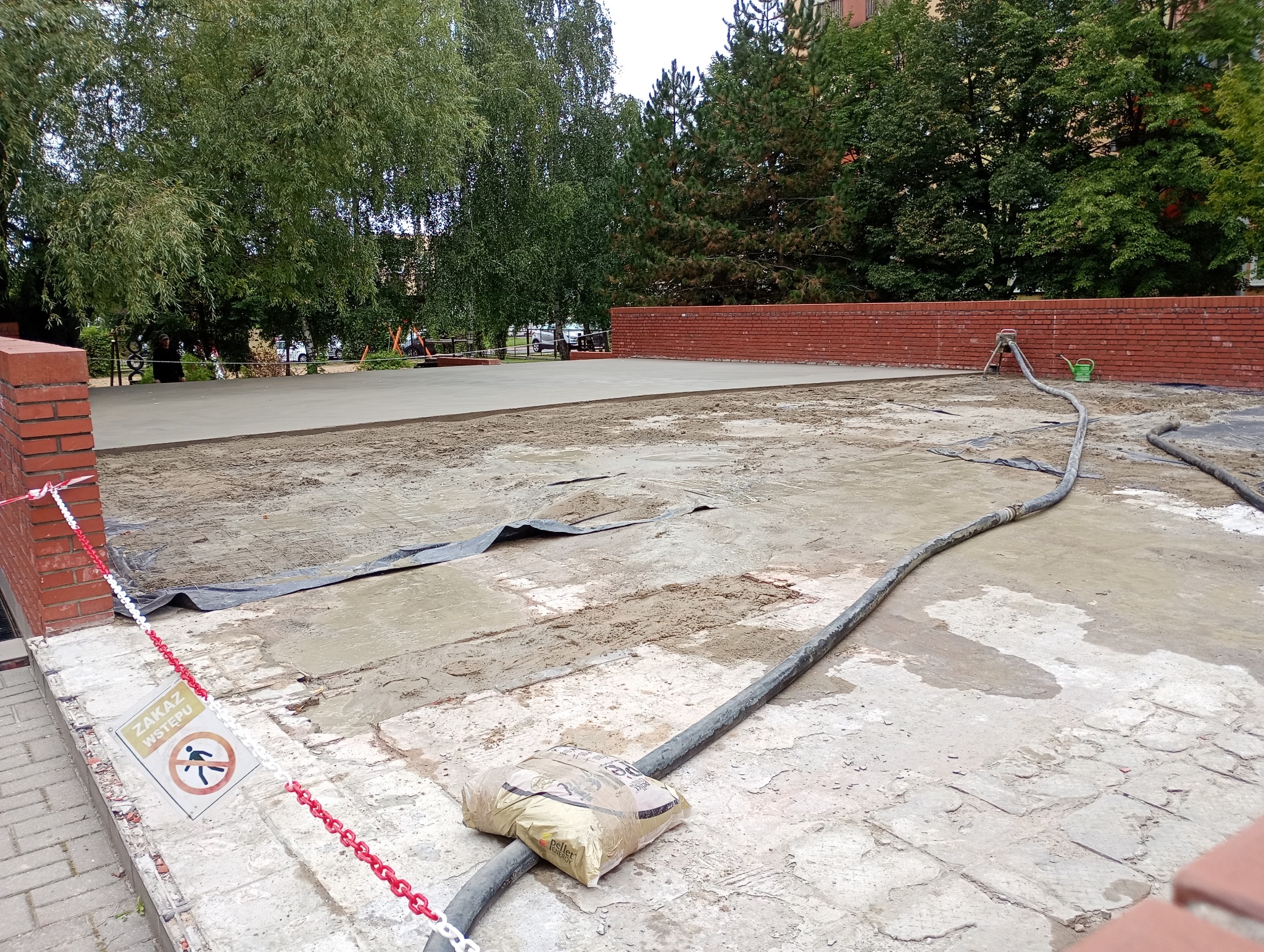 Wylewka jastrychowa w trakcie realizacji: świeża, szara powierzchnia, wąż do betonu, worek z zaprawą, znak zakazu wstępu, ceglany murek w tle, otoczenie zieleni miejskiej.