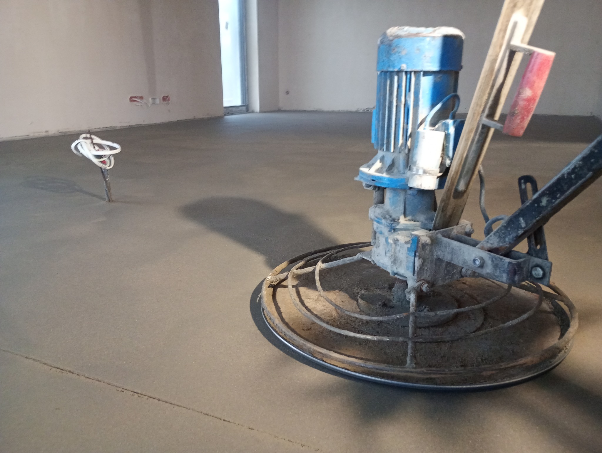 Zacieraczka mechaniczna do posadzek jastrychowych w trakcie pracy, widoczne przewody elektryczne wystające z podłoża. Szara, gładka powierzchnia świeżo wylanej posadzki.