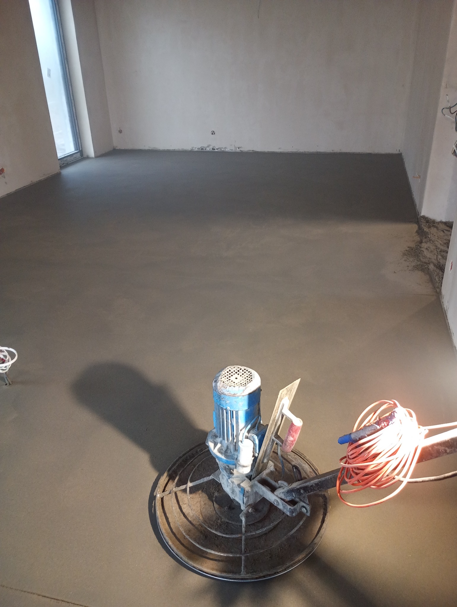 Wnętrze z maszyną do wygładzania posadzki jastrychowej, świeżo wylana, szara powierzchnia, widoczne ślady pracy, kabel zasilający, surowe ściany w tle.