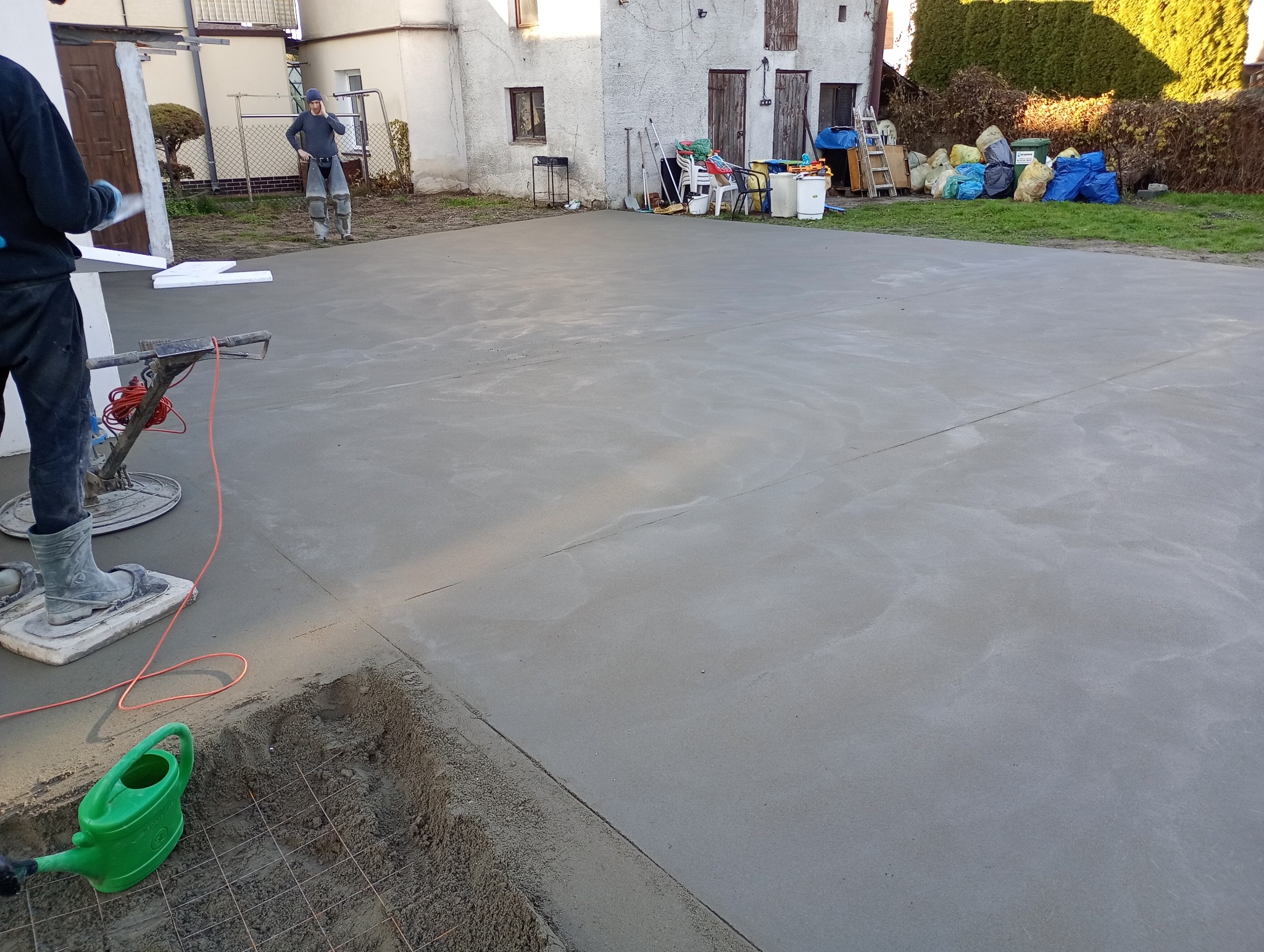 Świeżo wylana, gładka posadzka jastrychowa na zewnątrz, z widocznymi narzędziami i pracownikiem w tle. W rogu zielony konewka i siatka zbrojeniowa.