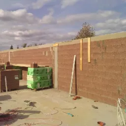 Wznoszenie ściany z bloczków ceramicznych z widocznymi elementami konstrukcyjnymi, ociepleniem i narzędziami na placu budowy w słoneczny dzień.