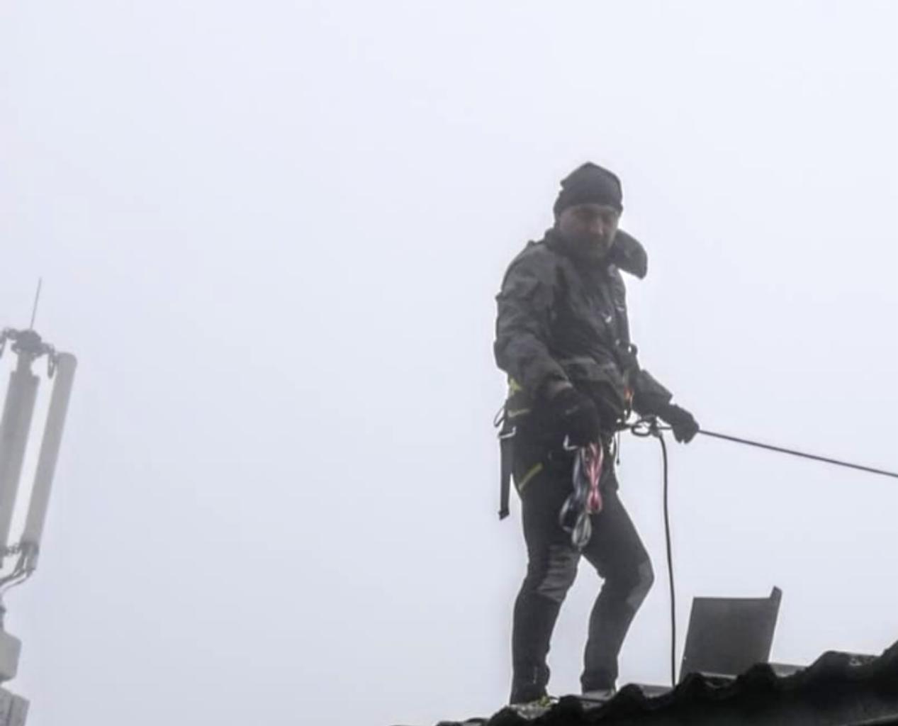 Pracownik w uprzęży na dachu w gęstej mgle, obok antena telekomunikacyjna.