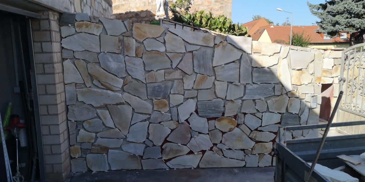 Ściana zewnętrzna wykończona nieregularnymi płytami kamiennymi w odcieniach beżu i szarości, przylegająca do ceglanego narożnika budynku, z widoczną bramą w tle.