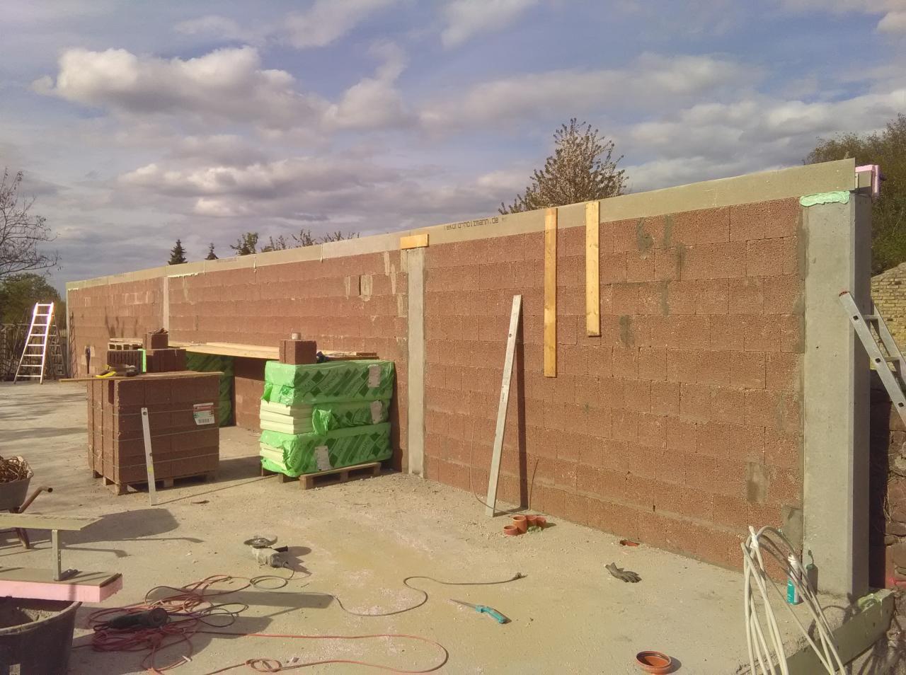 Wznoszenie ściany z bloczków ceramicznych z widocznymi elementami konstrukcyjnymi, ociepleniem i narzędziami na placu budowy w słoneczny dzień.