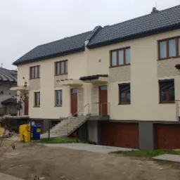 Nowo wybudowane segmenty mieszkalne z drewnianymi drzwiami wejściowymi i bramami garażowymi, widoczne schody z balustradami i teren budowy w tle.