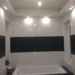 Nowoczesna łazienka z biało-czarnymi płytkami, podwieszanym sufitem z oświetleniem punktowym i nową wanną zabezpieczoną folią. Widoczne pudełka z akcesoriami do oświetlenia Polux.