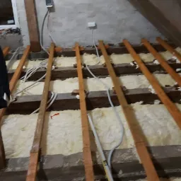 Poddasze w trakcie ocieplania: widoczne drewniane legary, izolacja z wełny mineralnej, przewody elektryczne oraz poziomica oparta o legar.