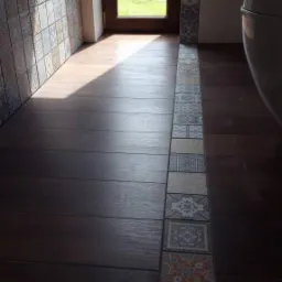 Ciemne drewnopodobne panele podłogowe w łazience, oddzielone od ściany pasem dekoracyjnych, wzorzystych płytek, z widokiem na zieleń przez okno.