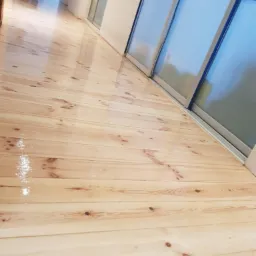 Świeżo polakierowana, jasna drewniana podłoga z widocznymi słojami, odbijająca światło, ułożona wzdłuż ściany z przesuwnymi drzwiami.