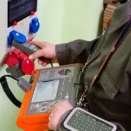 Mężczyzna w kombinezonie mierzy parametry elektryczne w rozdzielnicy za pomocą specjalistycznego miernika, widoczne kable pomiarowe i wyświetlacz urządzenia.