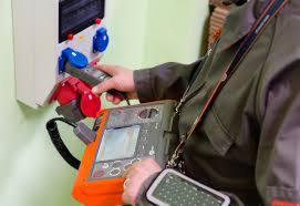 Mężczyzna w kombinezonie mierzy parametry elektryczne w rozdzielnicy za pomocą specjalistycznego miernika, widoczne kable pomiarowe i wyświetlacz urządzenia.