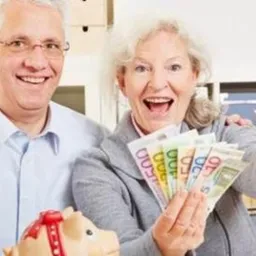 Uśmiechnięci seniorzy trzymający wachlarz banknotów euro i pokazujący kciuki w górę, obok świnki skarbonki z czerwoną kokardką.