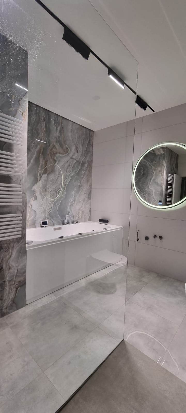 Nowoczesna łazienka z wanną z hydromasażem, szklaną kabiną prysznicową i okrągłym lustrem z podświetleniem LED. Marmurowa ściana i szara podłoga z płytek.
