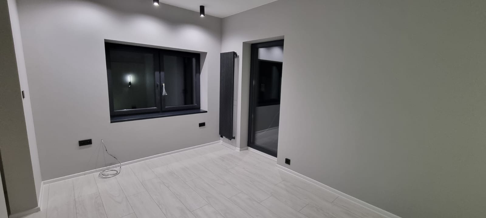 Szare, nowocześnie wykończone wnętrze pokoju z oknem w czarnej ramie, grzejnikiem panelowym i jasną podłogą imitującą drewno. Minimalistyczny design.