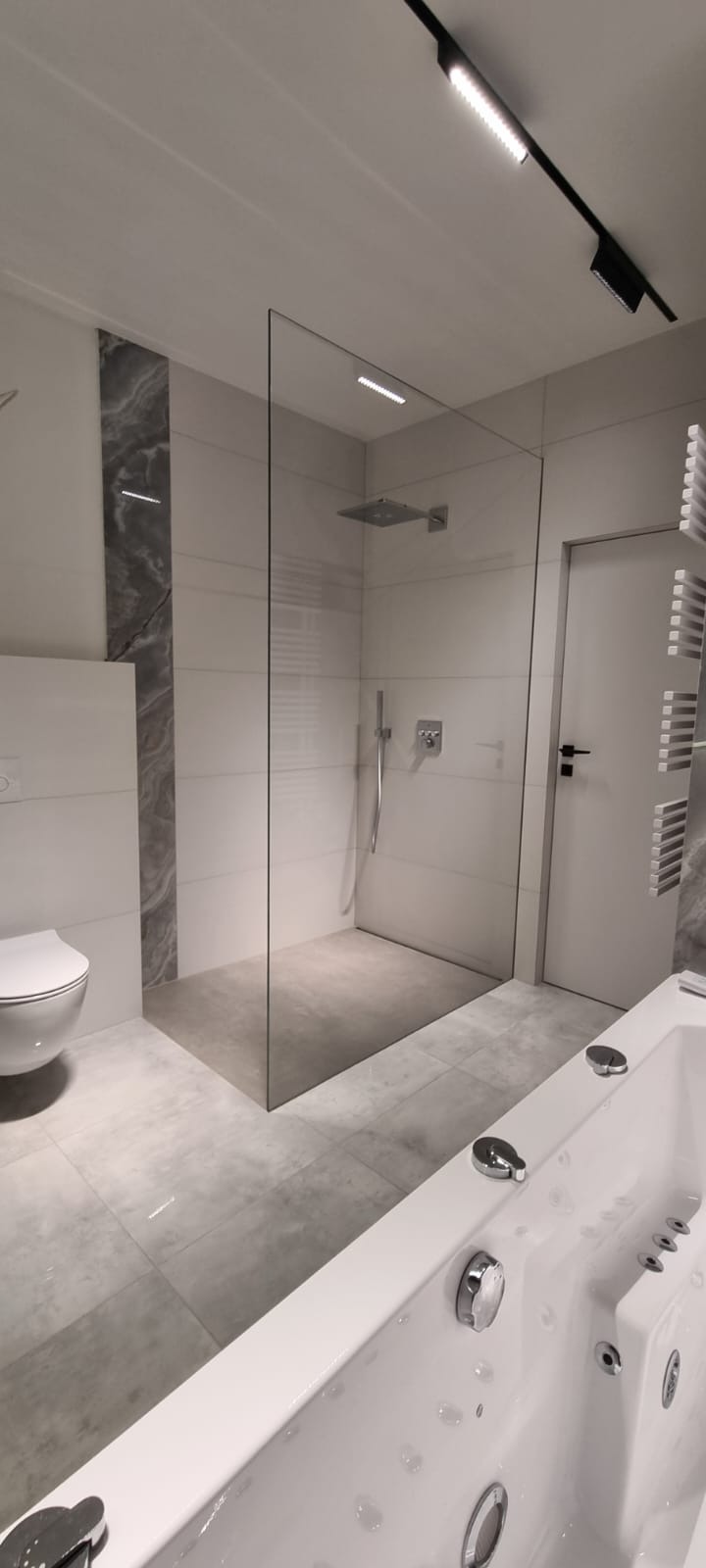 Nowoczesna łazienka z kabiną typu walk-in, białą armaturą i szarą płytką. Minimalistyczny design, oświetlenie punktowe i grzejnik drabinkowy. Wykończenie wnętrz.