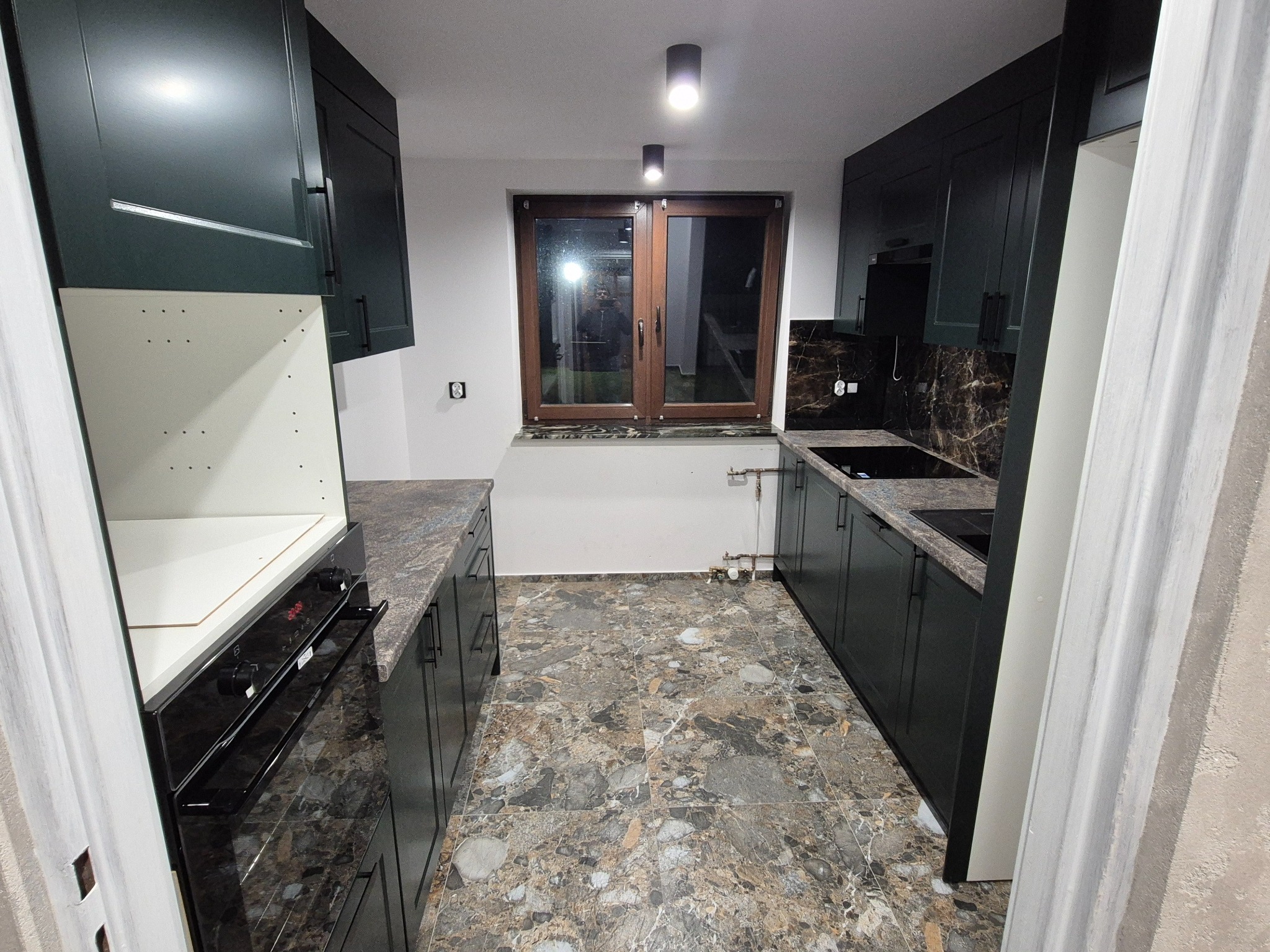 Nowoczesna kuchnia z ciemnymi szafkami, marmurowym blatem i podłogą. Widoczne okno i oświetlenie sufitowe. Częściowo widoczny piekarnik i fragment futryny drzwi.