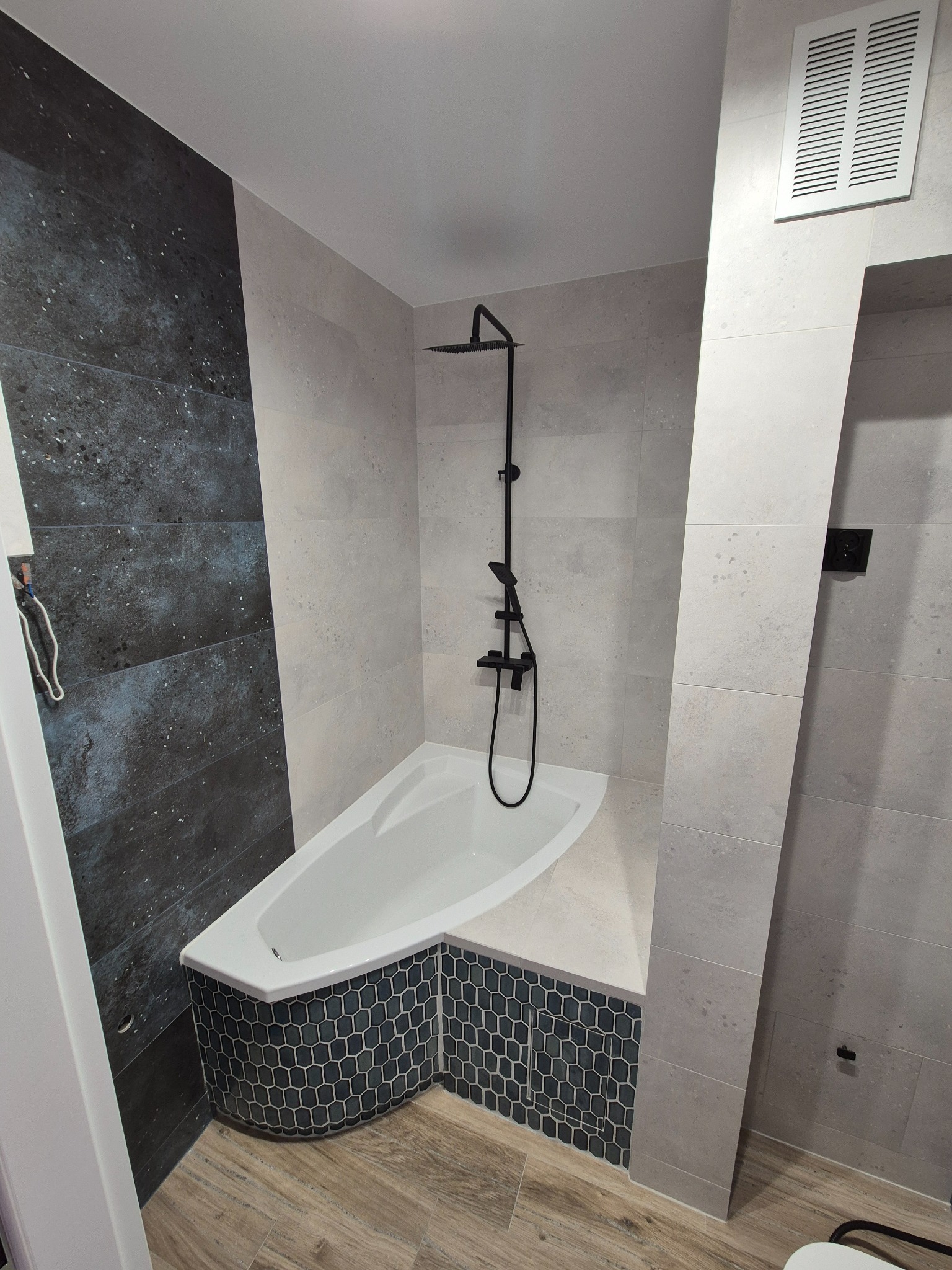 Nowoczesna łazienka po remoncie: asymetryczna wanna obudowana mozaiką, czarna armatura, szare płytki imitujące kamień i drewnopodobna podłoga. Minimalistyczny design.
