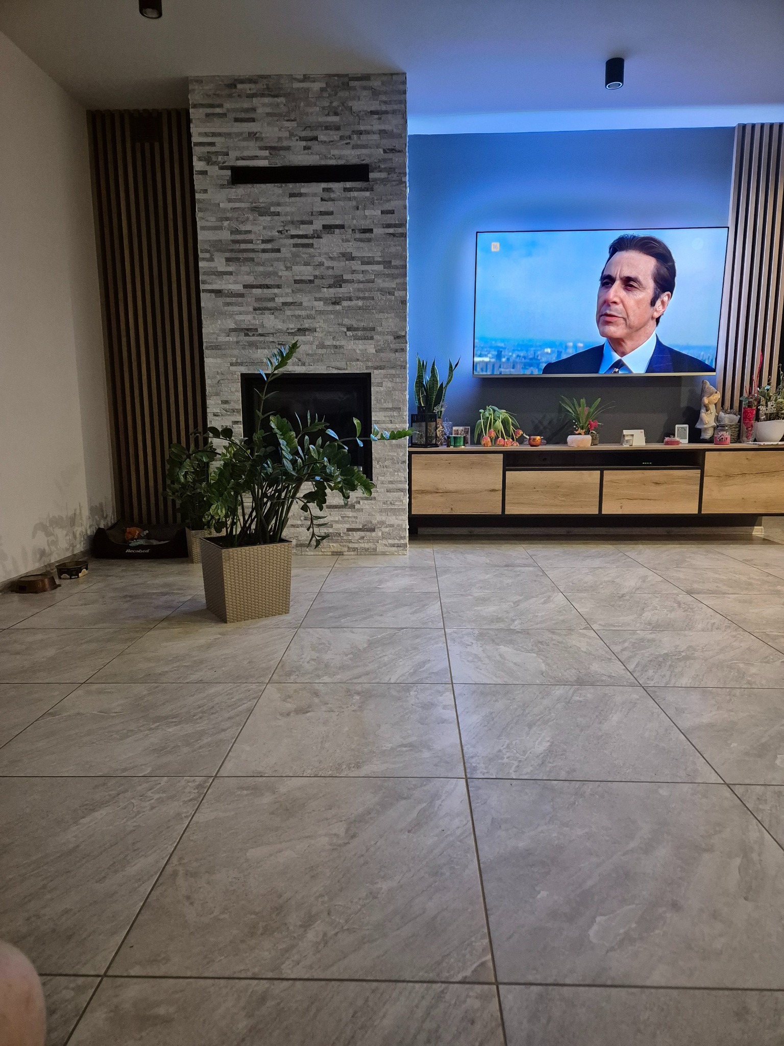 Salon z szarymi płytkami podłogowymi, kominkiem z cegły i telewizorem z włączonym filmem. Widoczne drewniane lamele na ścianie i roślina doniczkowa.