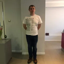 Mężczyzna w białej koszulce z czarnymi znakami, stojący na kafelkach z rękami założonymi za plecami, w jasnym pomieszczeniu.