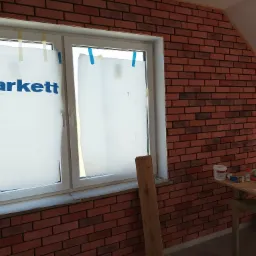 Ściana wewnętrzna wyłożona czerwoną cegłą klinkierową, okno zabezpieczone folią z logo Tarkett, widoczny stolik z narzędziami i materiałami budowlanymi.