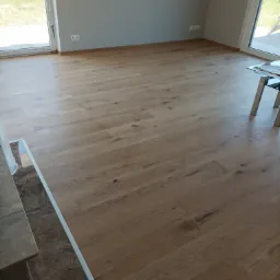 Jasny, dębowy parkiet ułożony w nowym, jasnym pomieszczeniu z dużymi oknami, widoczne listwy przypodłogowe i fragment marmurowego wykończenia.