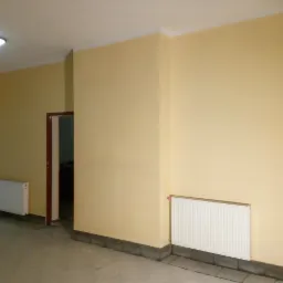 Wnętrze po malowaniu ścian na odcień żółci z widocznymi grzejnikami i fragmentem drabiny w lewym dolnym rogu, sugerujące prace wykończeniowe w trakcie.