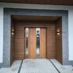 Nowoczesne wejście do domu z dwuskrzydłowymi drzwiami o drewnianej okleinie, otoczone szarą elewacją imitującą kamień i drewnianą podbitką dachową, oświetlone dwoma kinkietami.