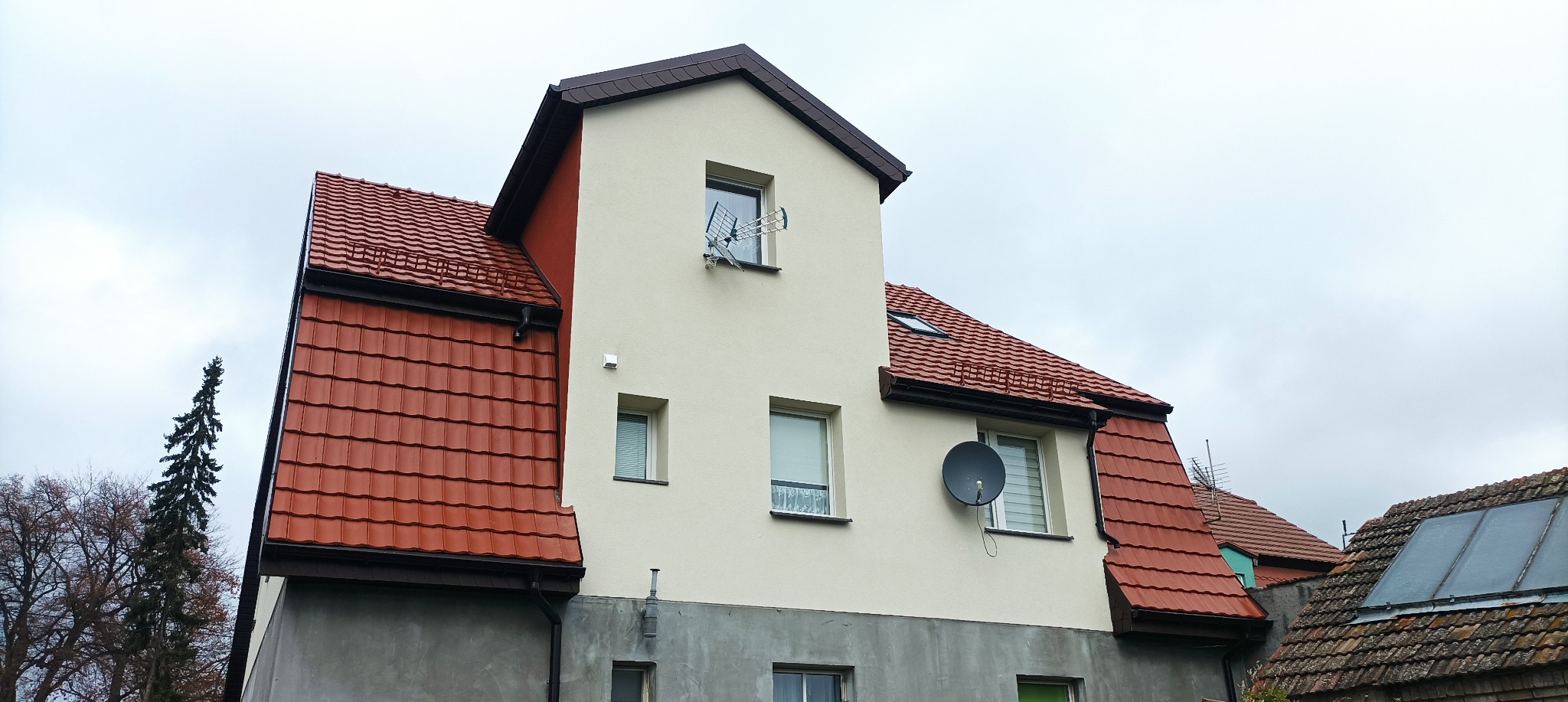 Dwukondygnacyjny budynek z nową, jasną elewacją w odcieniach beżu i szarości, z czerwonym dachem z dachówki, anteną satelitarną i anteną telewizyjną na ścianie, sfotografowany w pochmurny dzień.